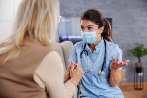 Friendly nurse with face mask sitting at home with senior woman and giving her advices