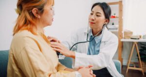 Asian woman doctor holding stethoscope examining with patient medical visit at home hospital.