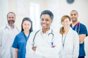 A group of doctors are inside a clinic. They are standing in a group and smiling for the camera and the camera focuses on the woman is the centre.