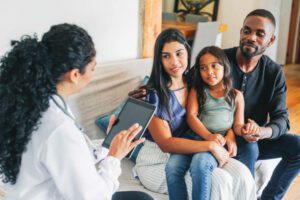 Young family sitting on sofa listening to the advice from a female doctor using digital tablet. Doctor on house call giving medical consultation to a young family.